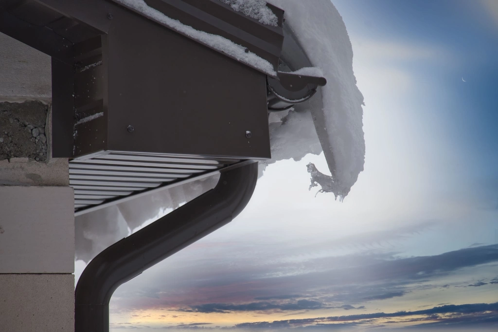 Thick snow and ice overhanging a roof edge beside a gutter and downspout